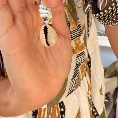 Pendente artesanal com búzio natural e contas brancas na mão de pessoa com roupa estampada.