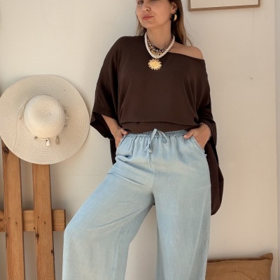 Mulher com blusa castanha escura, calças largas azul claro, colares e brincos dourados, chapéu bege pendurado, almofada bege, quadro minimalista numa parede branca.
