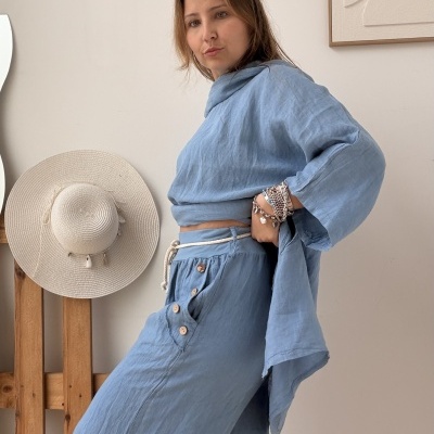 Conjunto de roupa azul claro com calças e camisa largas usadas por mulher em interior com chapéu de palha na parede