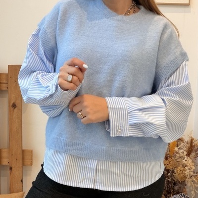 Mulher com camisola azul clara sobre camisa branca riscada azul e calças pretas num ambiente interior.