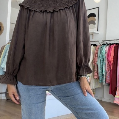 Blusa castanha com gola e punhos franzidos, jeans azuis claros e roupas ao fundo