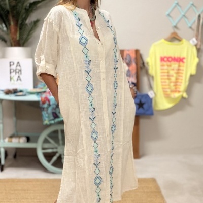 Vestido comprido branco com bordados azuis e sandálias pretas em ambiente interior.
