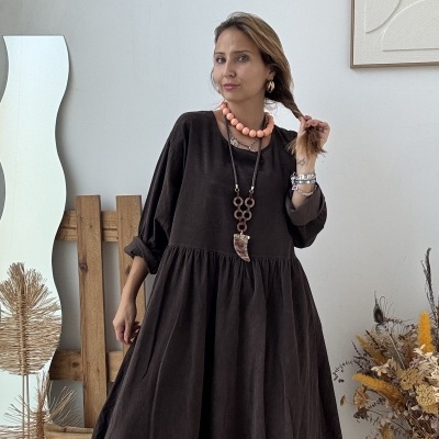 Mulher com vestido castanho escuro e colares em ambiente interior decorado.
