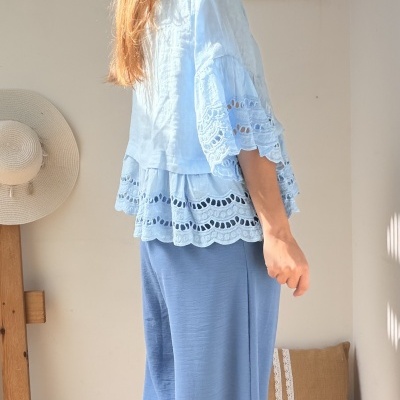 Mulher de perfil com blusa azul clara rendada e calças azuis largas em ambiente interior