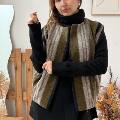 Mulher a usar camisola preta e colete riscado em tons de verde, cinza e azul com calças vinho, em quarto com decoração rústica.