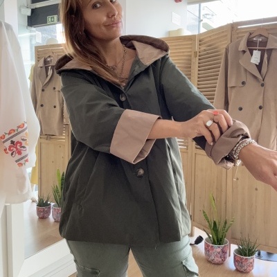 Casaco curto verde escuro com forro bege e botões, usado por mulher numa loja com divisórias e casacos ao fundo.