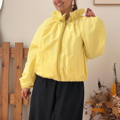 Mulher com casaco amarelo claro de fecho, calças pretas largas, em interior decorado com cadeira de madeira e vaso com flores secas