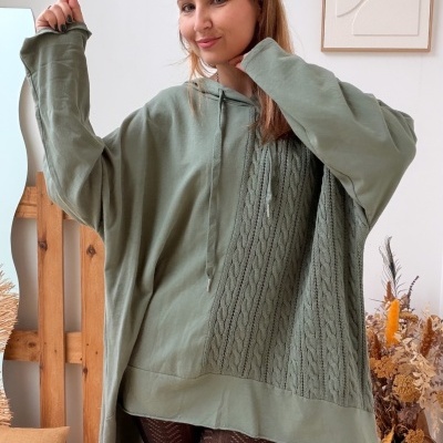 Mulher a usar camisola oversized verde com padrão knitted e capuz, meia preta e botas pretas no interior de uma casa decorada.