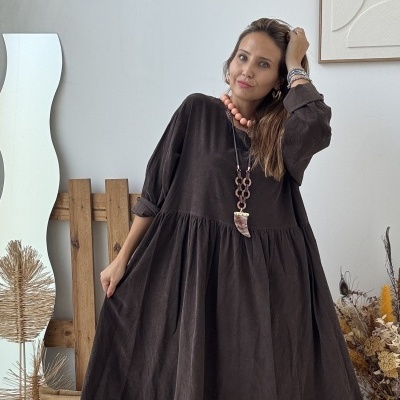 Vestido comprido castanho escuro com saia franzida e acessórios cor de salmão, mulher em ambiente com decoração natural.