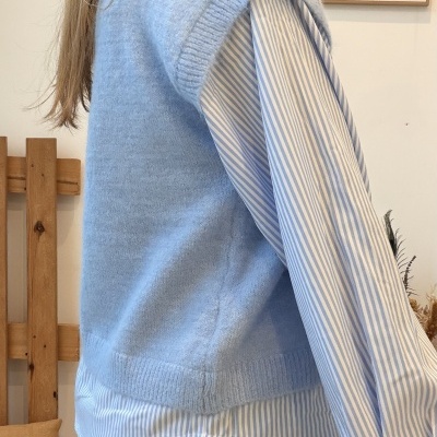 Pessoa a usar camisola de malha azul clara com camisa riscada azul e branca e calças pretas em interior