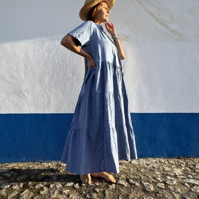 Vestido azul claro de algodão com corte evasê, manga curta, usado com chapéu de palha em fundo de parede branca e azul.