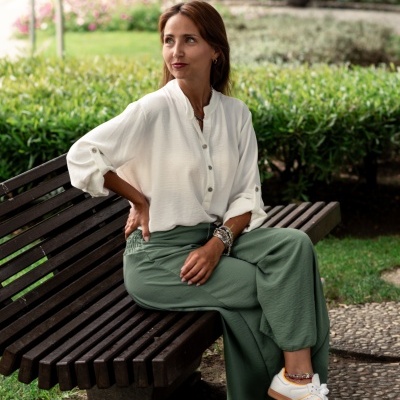 Mulher sentada num banco de madeira num parque com camisa branca e calças verdes largas