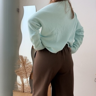 Pessoa de costas com camisola azul claro e calças castanhas num interior com chão de madeira e espelho