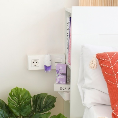 Ambientador elétrico lilás e branco junto a cama branca com almofada vermelha e plantas verdes