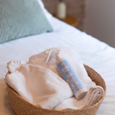 Cesta de fibra natural com toalhas brancas e frasco azul com pulverizador numa cama branca.