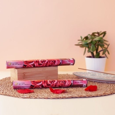 Dois pacotes de incenso de rosas com embalagem rosa e imagens de rosas sobre superfície de ráfia com pétalas e planta ao fundo