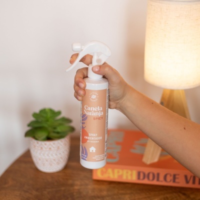 Spray ambientador Canela Naranja segurado por uma mão ao lado de planta e lâmpada