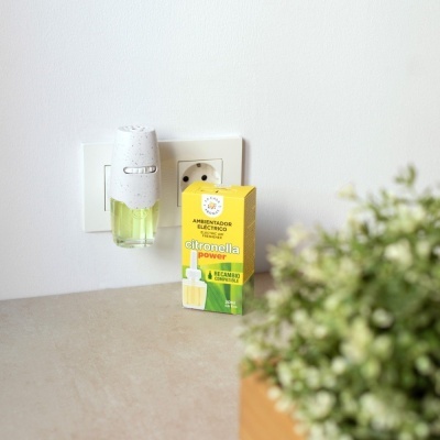 Ambientador elétrico citronella power ligado a tomada branca com embalagem de recarga amarelo verde e planta desenfocada