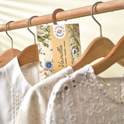 Embalagem de vela perfumada Vanilha pendurada num cabide entre roupa branca texturizada