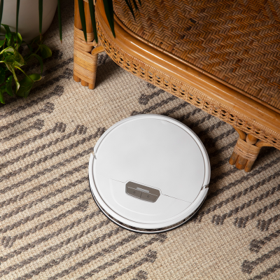 aspirador robô branco circular no tapete, próximo a planta e móvel de rattan