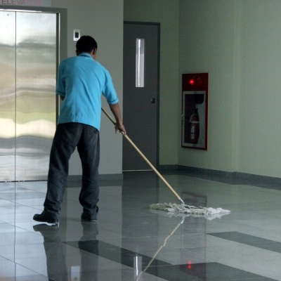 Homem a limpar chão com esfregão numa zona interior com elevador e extintor