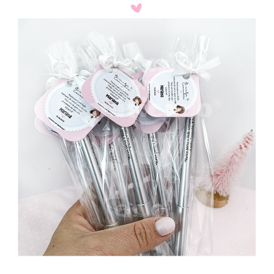 Cinco canetas prateadas embaladas individualmente com etiquetas cor-de-rosa e branco, seguradas por uma mão com unhas brancas sobre superfície rosa clara com decoração.