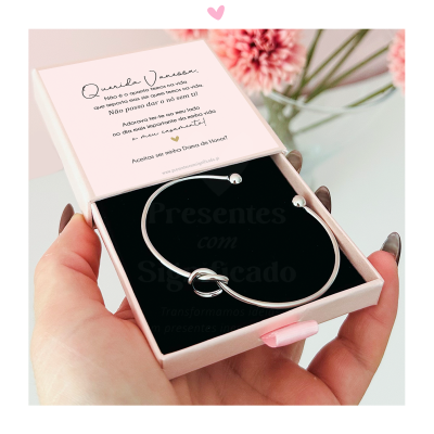 Pulseira prateada em caixa rosa com mensagem personalizada e flores rosas ao fundo