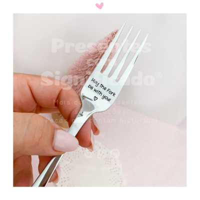 Garfo de aço inoxidável com frase gravada segurado numa mão com fundo rosa e branca