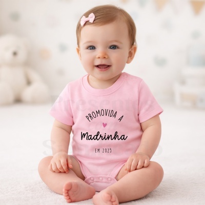 Bebé de body rosa com texto PROMOVIDA A Madrinha EM 2025 sentada com laço na cabeça