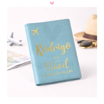 Capa de passaporte azul claro com texto dourado e decoração de avião, chapéu de palha e óculos de sol ao fundo