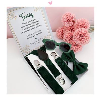 Conjunto verde com suspensórios, laço, óculos de sol e cartão em caixa branca, flores rosa ao lado.