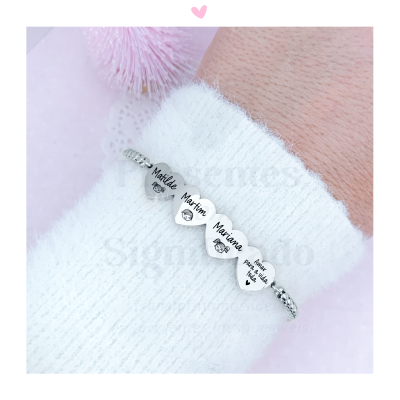 Pulseira prateada com medalhas em forma de coração com nomes e desenhos, usada num pulso com manga branca de malha.
