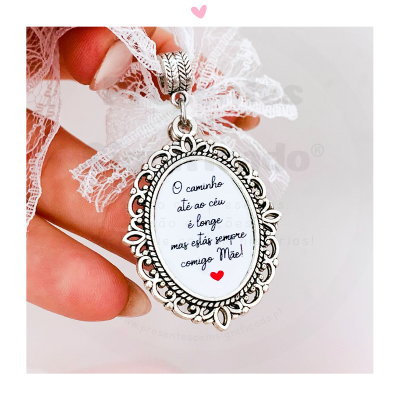Pingente prateado com mensagem para mãe em português, segurado por uma mão com unhas nude e renda branca ao fundo.
