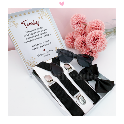 Conjunto preto para menino com gravata borboleta, suspensórios e óculos de sol numa caixa com mensagem em português e flores cor-de-rosa ao fundo