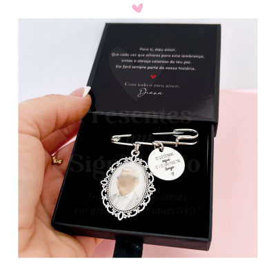 Alfinete de segurança prateado com medalha oval com foto desfocada e pendente redondo com texto, numa caixa preta.