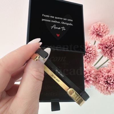 Pulseira dourada com detalhes pretos numa caixa preta com mensagem romântica e flores cor-de-rosa ao fundo