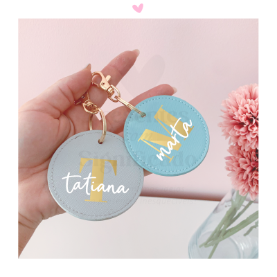 Porta-chaves redondos em couro sintético com iniciais e nomes personalizados à mão, segurados por uma mão, fundo rosa e flores cor-de-rosa à direita