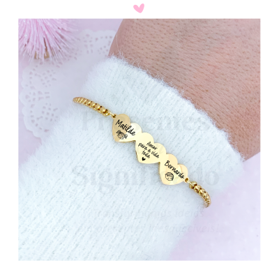 Pulseira dourada com três corações gravados com nomes, no pulso com camisola branca