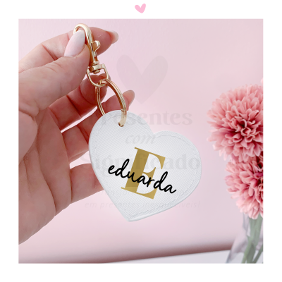 Chaveiro coração branco com nome eduarda e letra E dourada, mão segurando, fundo rosa com flores.