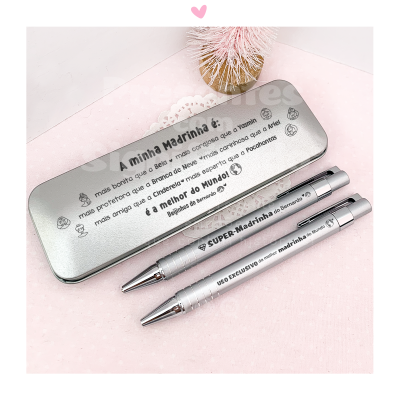 Estojo metálico prateado com texto e duas canetas também prateadas em superfície rosa clara com vaso de flores