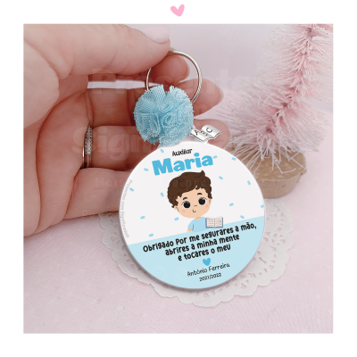 Chaveiro redondo branco com desenho de criança e texto, pompom azul e anel metálico, sobre base rosa com objeto rosa desenhado