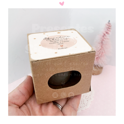 Caixa de vela pequena em cartão castanho com texto e estrelas douradas, ao lado árvore de Natal rosa