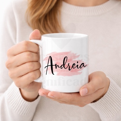 Caneca branca com o nome Andreia sobre fundo rosado, segurada por uma pessoa com suéter bege.