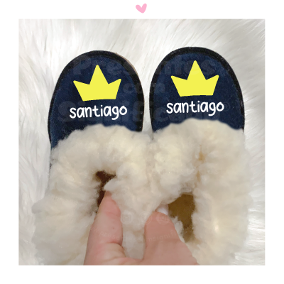 Chinelos azuis escuros com interior de pele de carneiro branco, coroa amarela e texto 'santiago'