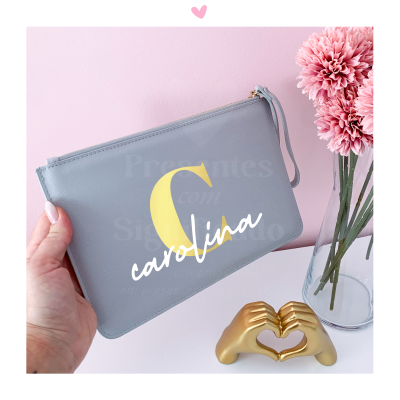 Carteira cinzenta com letra C amarela e nome Carolina em branco perto de flores rosas e escultura dourada em forma de mãos