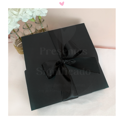 Caixa de presente preta com fita preta numa superfície branca com frascos de vidro e flores brancas ao fundo.