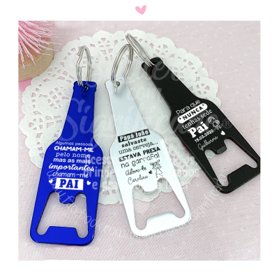 Três abridores de garrafa chaveiros azul, branco e preto com mensagens para o pai colocados sobre guardanapo branco rendado