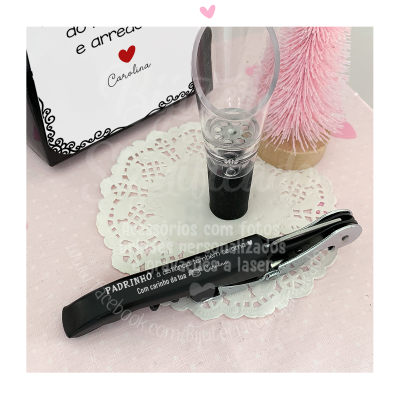 Conjunto de saca-rolhas preto e derramador transparente com base preta sobre renda branca, árvore decorativa rosa e caixa branca com texto.