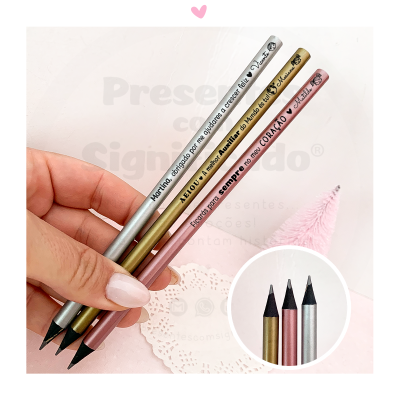 Três lápis de carvão metálicos com texto em português, numa mão, fundo branco e rosa claro