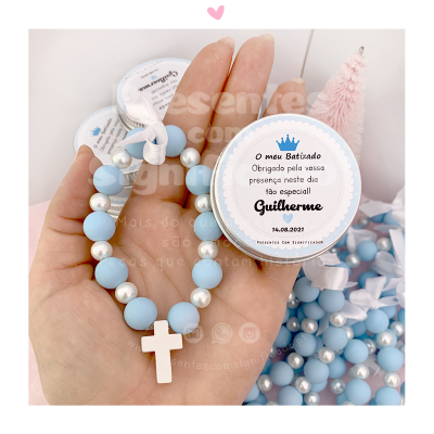 Pulseira azul e branca com crucifixo e embalagem para batizado com texto em português.